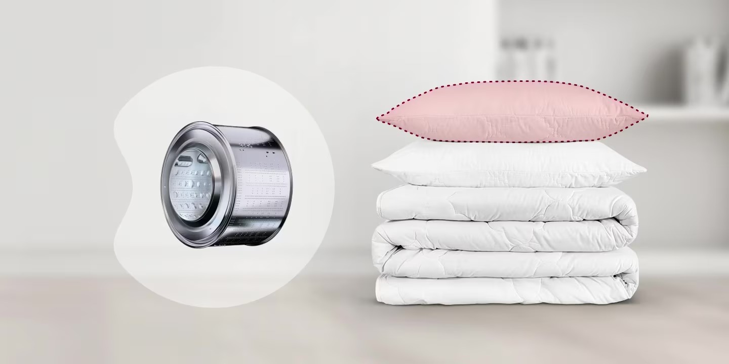 Gối màu hồng và chăn ga trắng xếp chồng bên cạnh lồng giặt kim loại của máy giặt